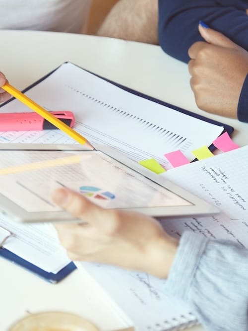 Hands reviewing printed progress reports and a tablet chart, marked with highlighter and sticky tabs