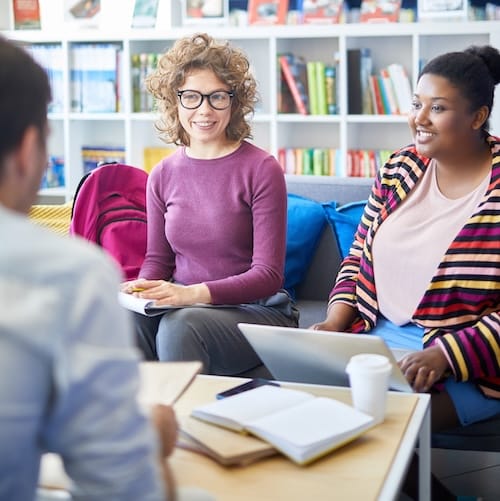 Three adults meeting around a low table in a school library, with open notebooks and a laptop